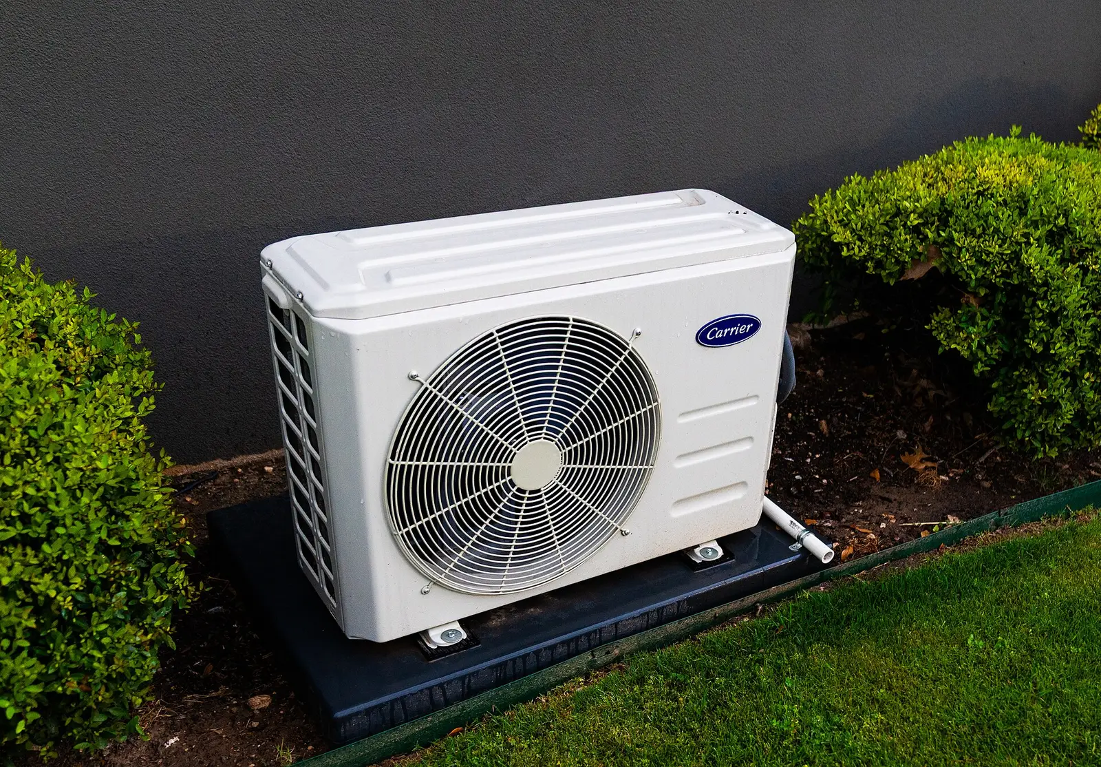 Petite unité extérieure de climatisation réversible posée sur un support au sol, entre deux haies