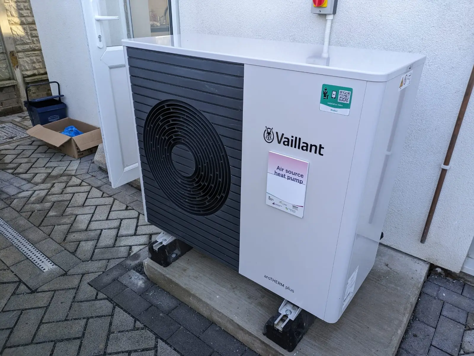 Unité extérieure blanche d’une pompe à chaleur air-eau posée au pied d’une façade, sur un dallage en chevrons